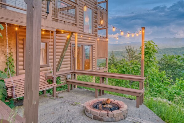A wooden cabin with a deck, a fire pit, and string lights.