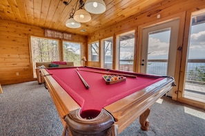 A pool table with a red felt surface, a cue stick, and a set of balls in a corner of a room with wooden walls and large windows.