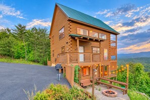 A log cabin with a porch, a fire pit, and a view of the surrounding forest.