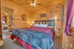 A wooden cabin bedroom with a bed, a ceiling fan, a mirror, and a nightstand with a lamp.