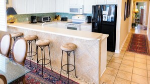 Una cocina moderna con armarios blancos, un refrigerador negro, un microondas y una cafetera. Hay tres taburetes y una mesa con cubierta de vidrio.