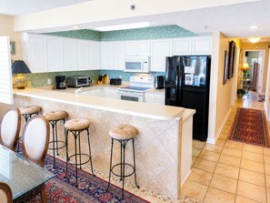 A modern kitchen with white cabinets, a black refrigerator, a microwave, and a coffee maker. There are three stools and a glass-top table. - Experience Beach Club III 1E! Oceanfront Paradise Awaits: Spacious 3-Bedroom Condo with Stunning Views! (North Myrtle Beach)