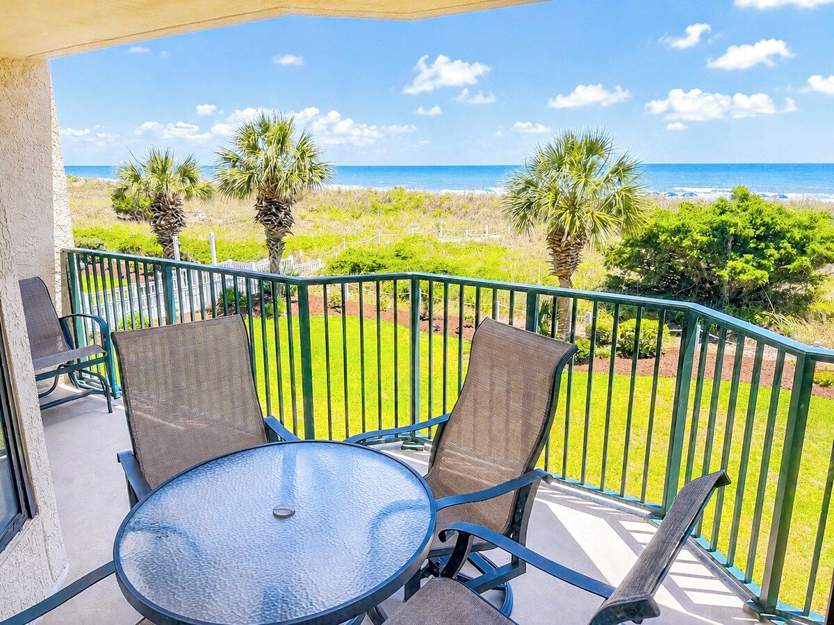 A balcony with a glass table and two chairs, overlooking a beach and grassy area.