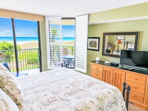 A bedroom with a bed, a dresser, a television, and a balcony with a view of the beach.