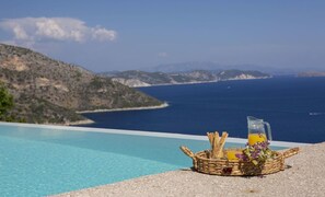 Uma cesta de vime com petiscos e uma bebida num terraço com vista para um mar azul e um cenário montanhoso.