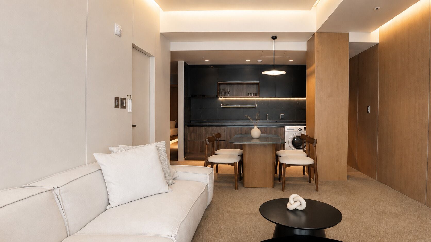 A modern living room with a sofa, a dining table, and chairs, a kitchen area with a washing machine, and a small decorative item on the floor.