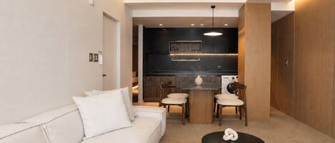 A modern living room with a sofa, a dining table, and chairs, a kitchen area with a washing machine, and a small decorative item on the floor.