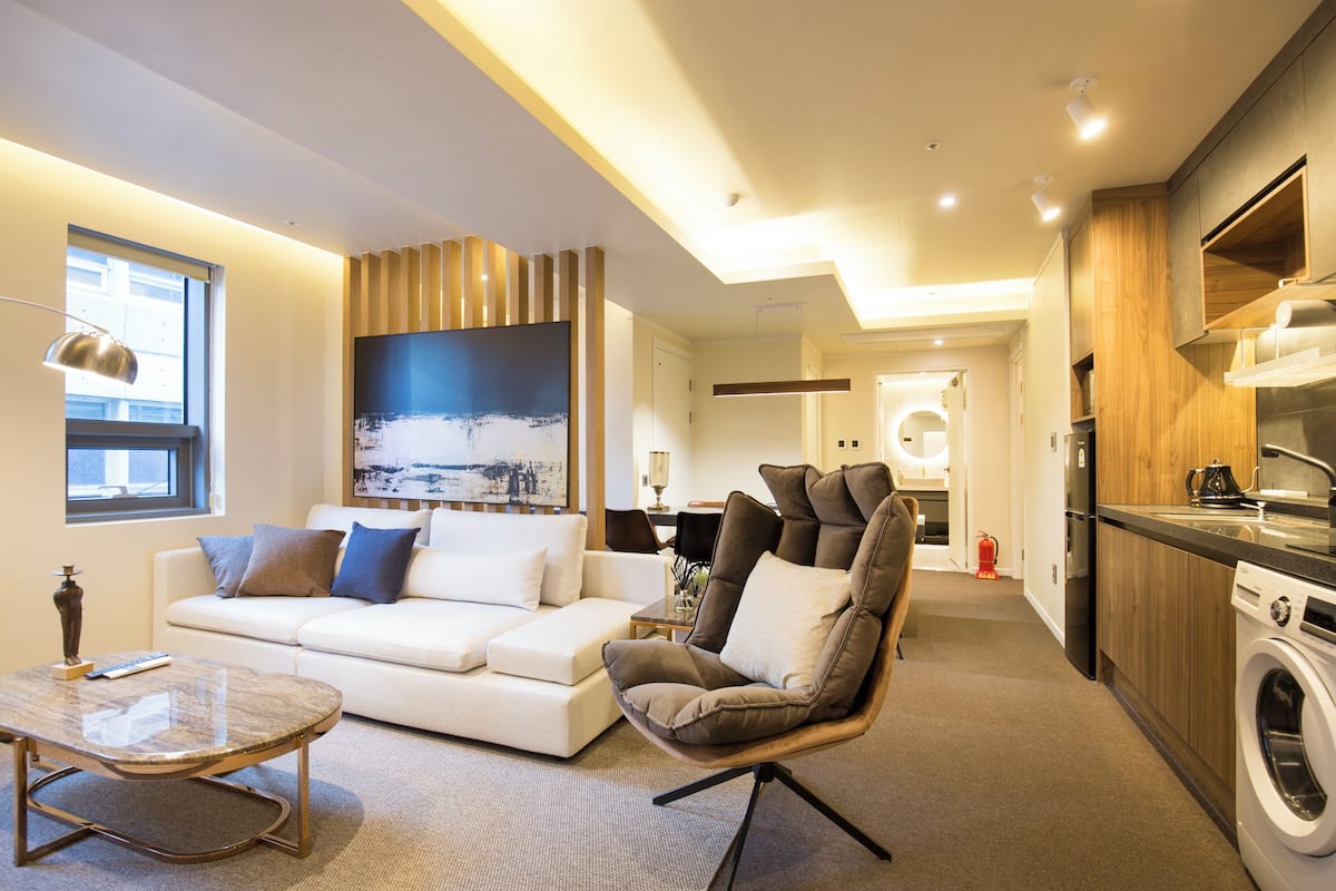 Ein modernes Wohnzimmer mit Sofa, Sessel und Couchtisch. Es gibt eine Küchenzeile mit Waschmaschine sowie einen Essbereich mit Tisch und Stühlen.