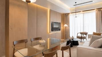 A modern hotel room with a glass dining table, wooden chairs, a sofa, and a TV mounted on the wall.
