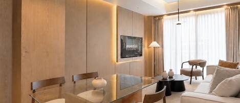 Una habitación de hotel moderna con una mesa de comedor de vidrio, sillas de madera, un sofá y un televisor montado en la pared.