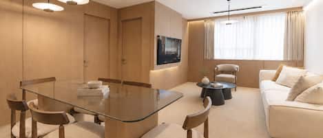 A modern living room with a glass dining table, wooden chairs, a sofa, and a TV mounted on the wall.
