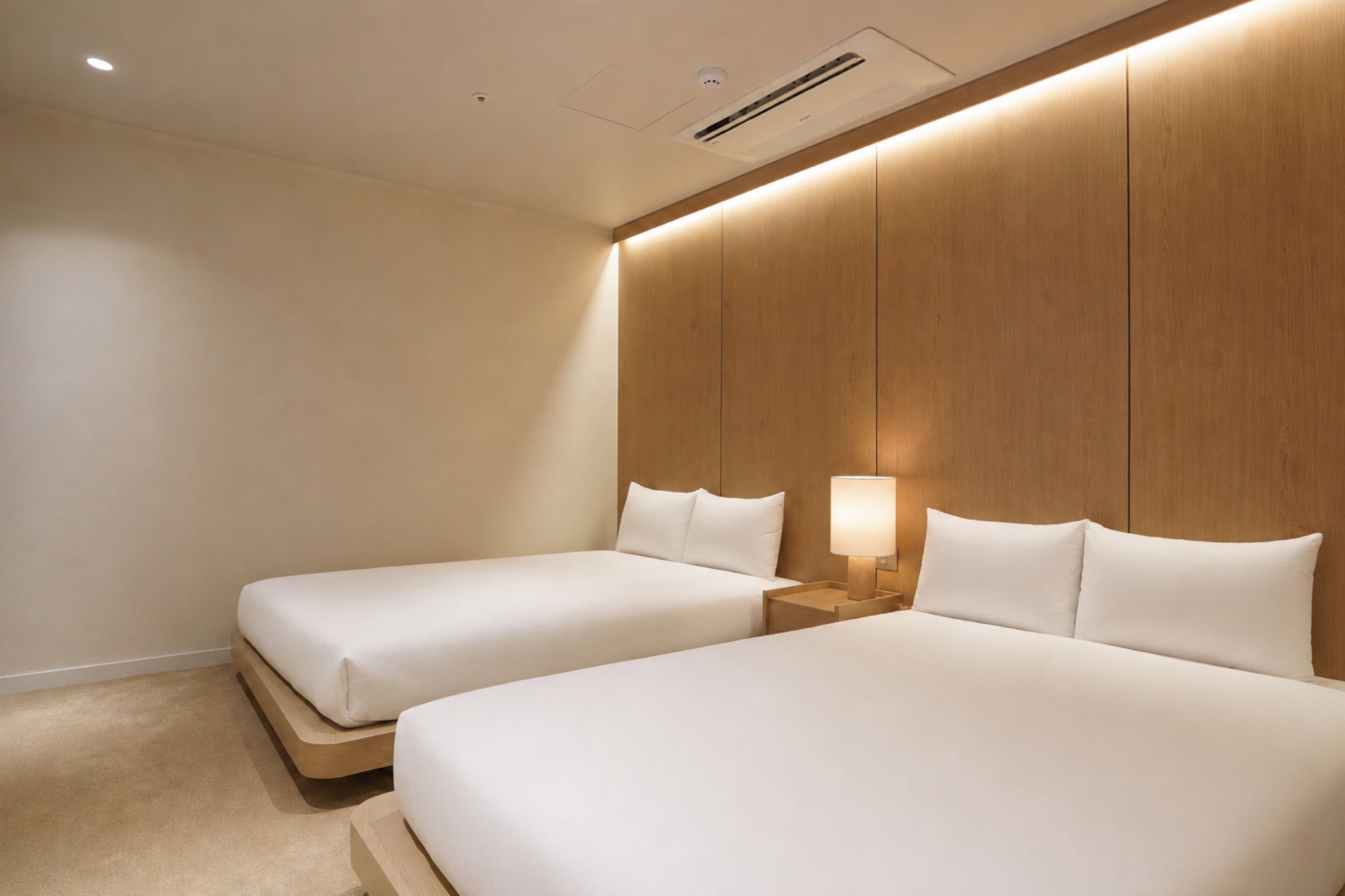 Habitación de hotel con dos camas individuales, cabecera de madera, mesita de noche con lámpara y rejilla de ventilación en el techo.
