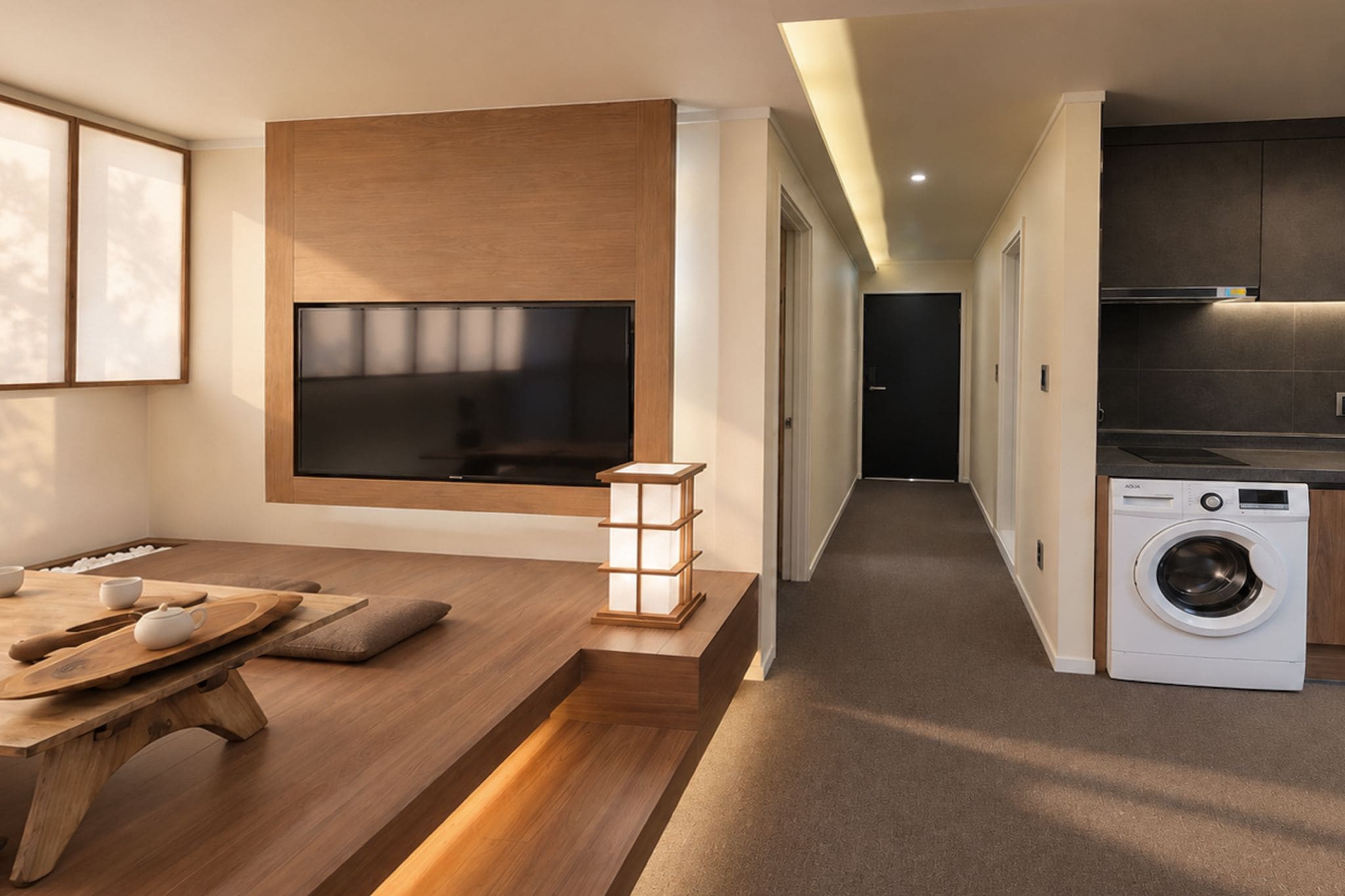A modern living room with a flat-screen TV, wooden furniture, and a kitchen area with a washing machine.