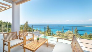 Un balcón con mobiliario de madera, barandilla de cristal y vistas al mar y las montañas.