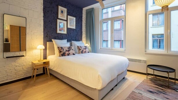 A modern bedroom with a large bed, a ceiling fan, a window with curtains, and a small table. - The Blue Parrot by SHWAY (Antwerp)