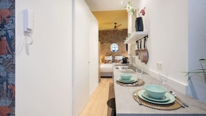 A narrow kitchen with a wooden floor, a white wall, and a brick wall in the background. There is a sink, a stove, and a shelf with kitchenware. - The Red Monkey by SHWAY (Antwerpen)