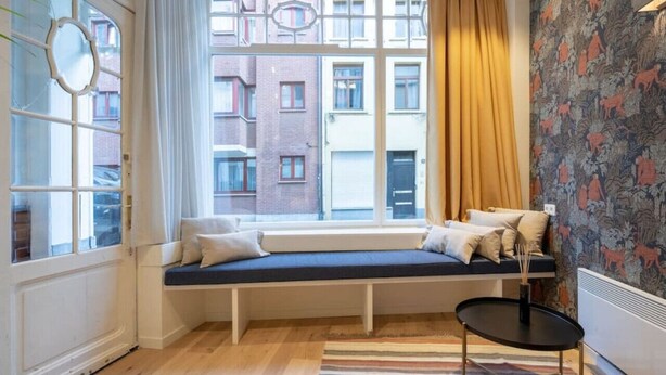A room with a built-in bench by a large window, a small round table, and a patterned wall. - The Red Monkey by SHWAY (Antwerpen)