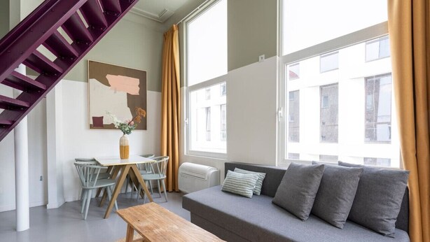 En moderne stue med en grå sofa, et træbord, et spisebord med stole, en lilla trappe og store vinduer med gardiner.