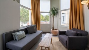 En moderne stue med en grå sofa, en mindre grå lænestol, et træbord og et stort vindue med gardiner.
