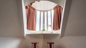 A room with a curved ceiling, a window with curtains, a desk with a lamp, and two stools. - The Suit by SHWAY Antwerp (Antwerpen)