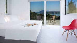 A bedroom with a bed, a red chair, and a view of a lake and mountains through a glass door. - Villa Anticlea (Lefkada)