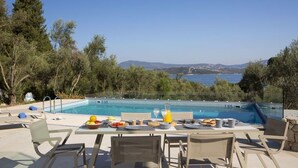 A poolside dining area with a table set for breakfast, overlooking a pool and a scenic view of hills and water. - Villa Anticlea (Lefkada)