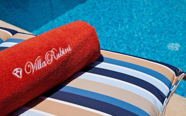 A red Villa Rubini towel on a striped lounge chair by a pool.