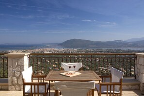 Una mesa de madera con mantel blanco, dos sillas y una copa de vino tinto en un balcón con vistas a una ciudad costera y montañas al fondo.