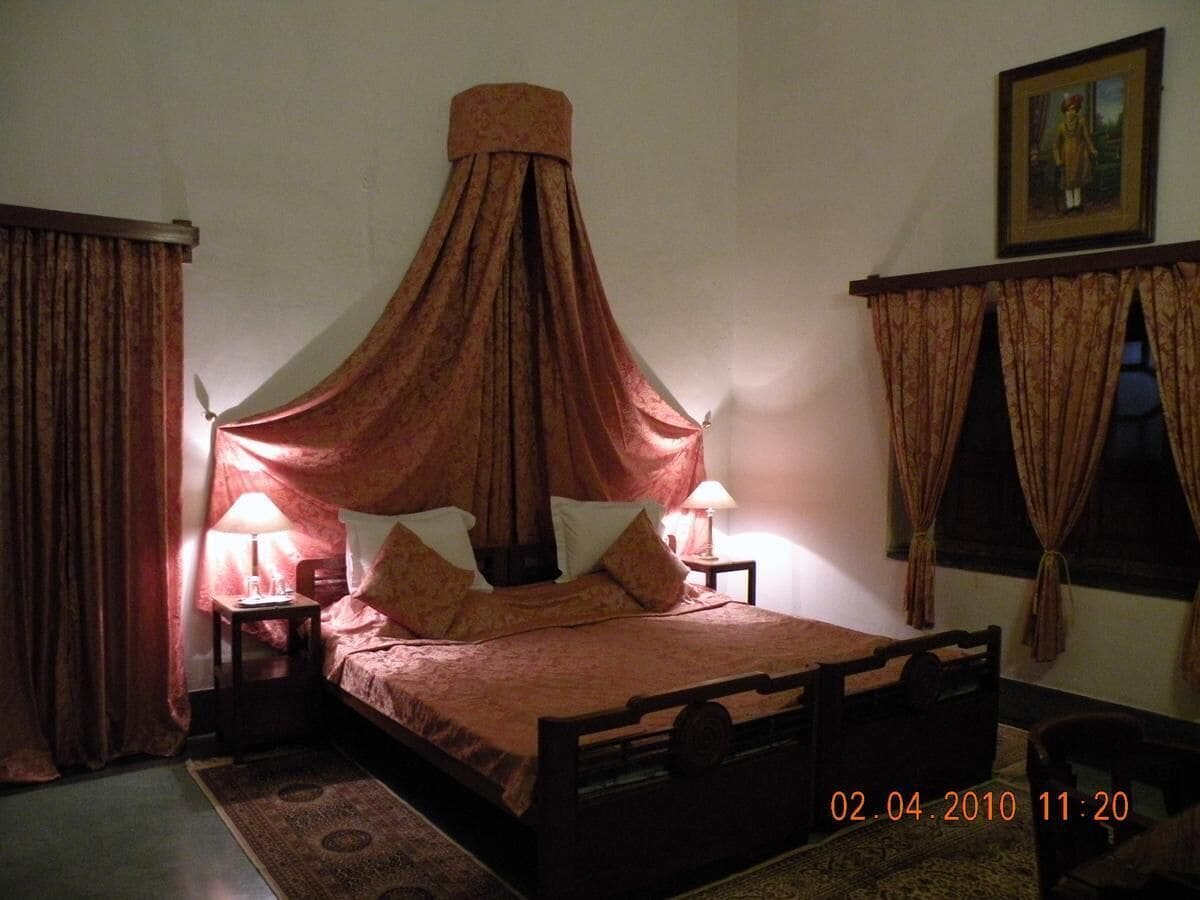 A room with a bed, two bedside tables, a nightstand, a window with curtains, and a painting on the wall.