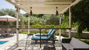 A covered outdoor seating area with lounge chairs, a table, and a pool. - Villa Niriides (Lefkada)