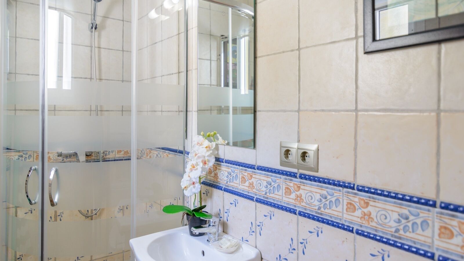 A bathroom with a glass shower enclosure, a white sink with a mirror above it, and decorative blue and orange tiles on the walls.