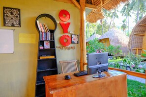 A wooden desk with a computer, keyboard, and mouse in a room with a thatched roof and a view of a tropical garden. - OYO 2039 Cici's Bungalow (Senggigi)