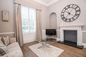 A living room with a sofa, a glass coffee table, a large wall clock, a fireplace, and a television.