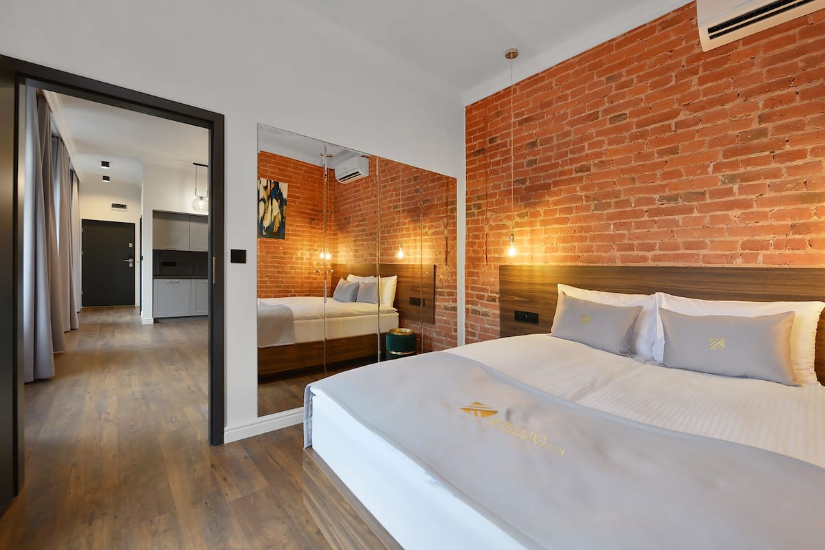 Ein modernes Hotelzimmer mit Backsteinwand, einem großen Bett mit weißer Bettwäsche und einer Glastrennwand, die das Schlafzimmer vom Wohnbereich abteilt.