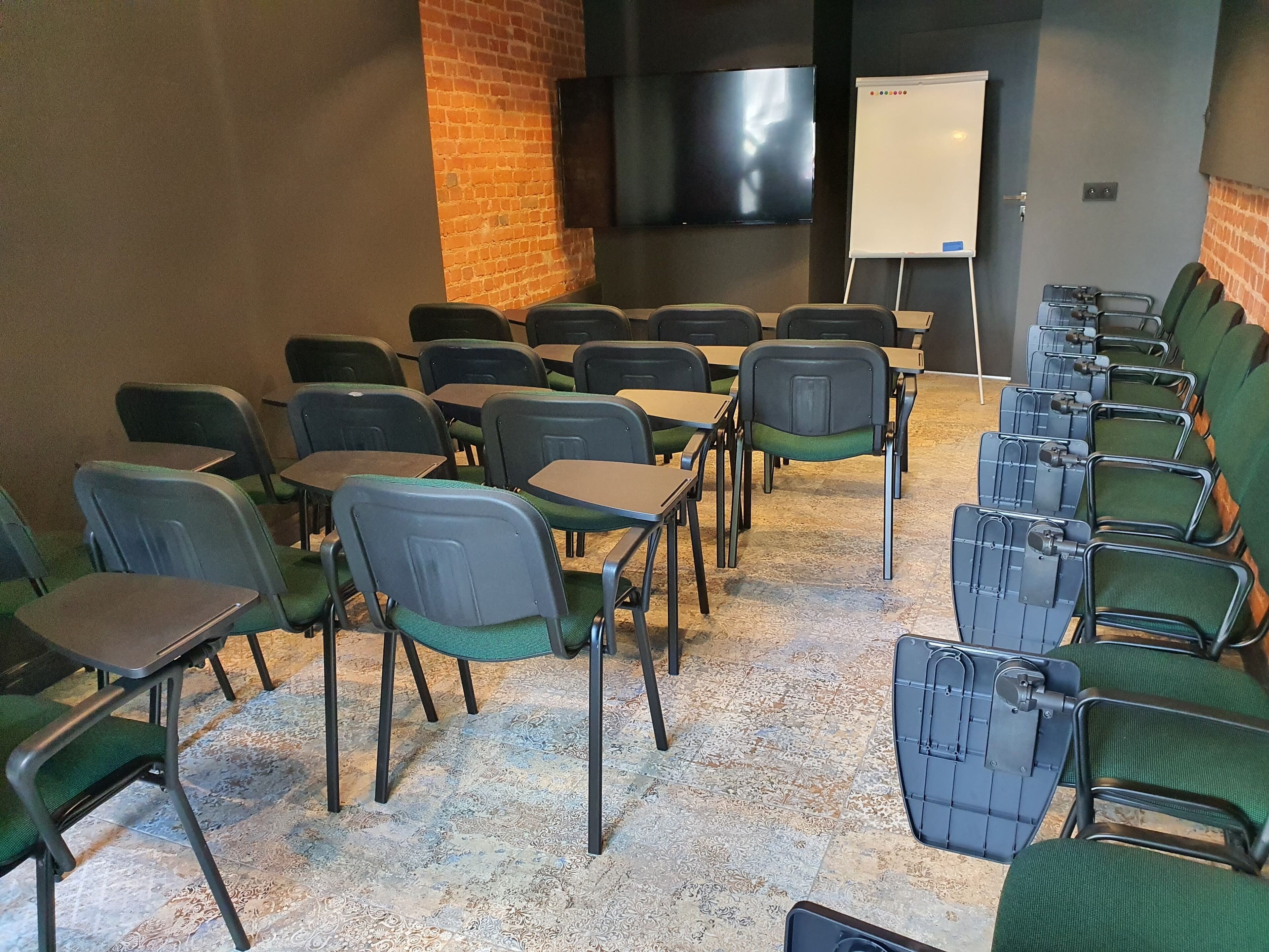 Un salón de conferencias con filas de sillas y mesas, una pizarra y un televisor en la pared.