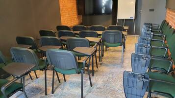 Un salón de conferencias con filas de sillas y mesas, una pizarra y un televisor en la pared.