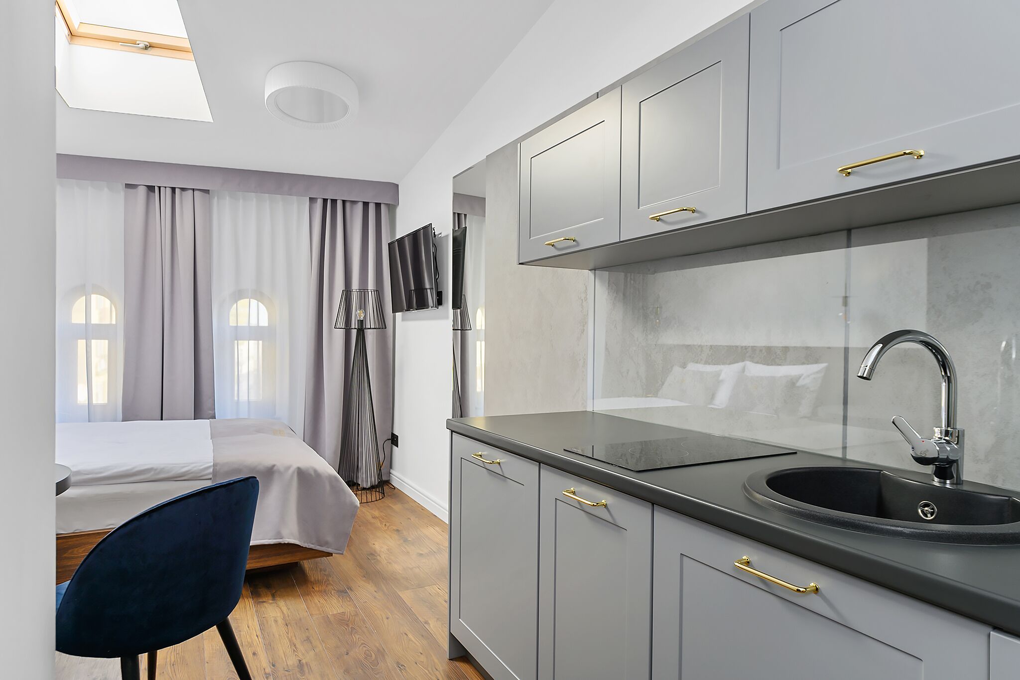 A modern kitchen with grey cabinets, a black countertop, and a sink. There is a bed with grey curtains and a blue chair in the adjacent room.