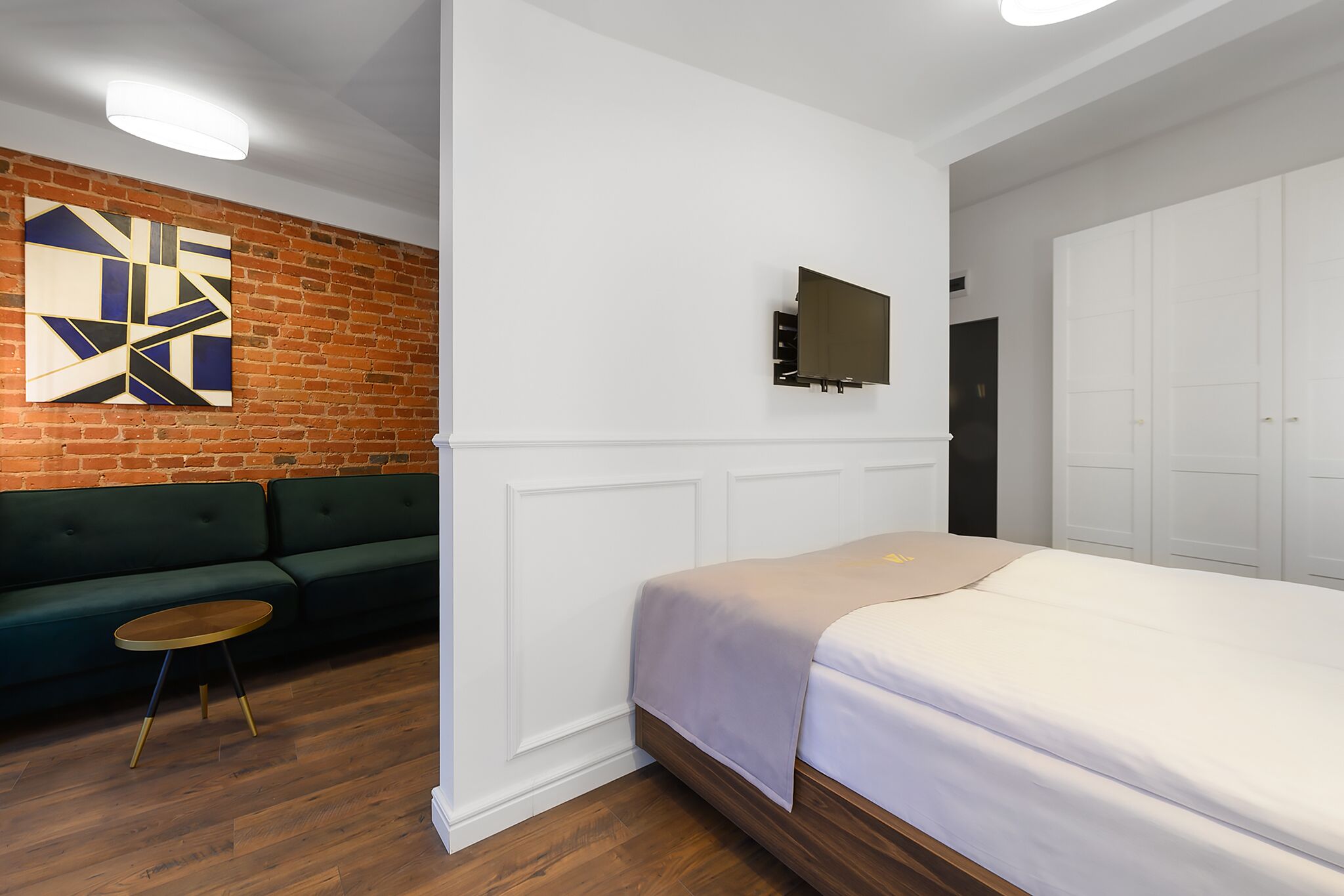Una habitación de hotel moderna con pared de ladrillo decorativa, un sofá verde, piso de madera, una cama con colcha blanca y un televisor de pantalla plana montado en la pared.