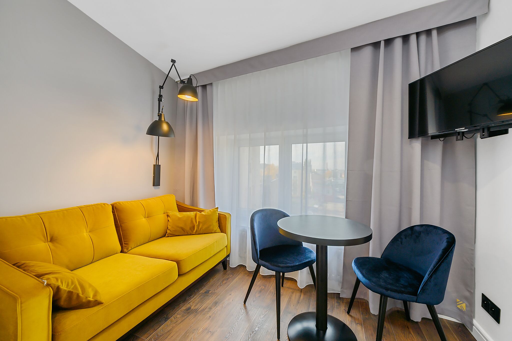 Un salon moderne avec un canapé jaune, deux fauteuils bleus, une table ronde, un téléviseur noir fixé au mur et des rideaux légers.