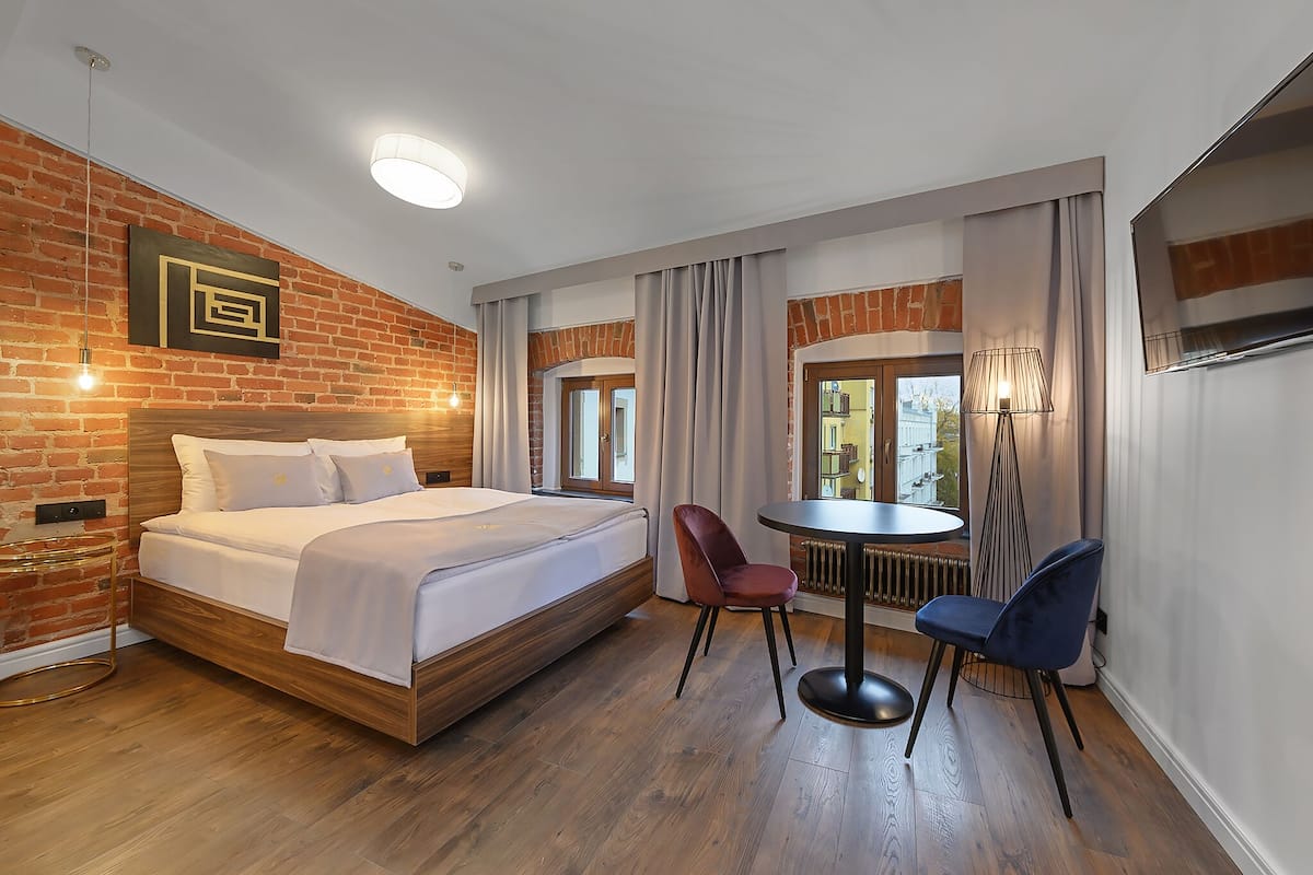 Ein Hotelzimmer mit Backsteinwand, einem hölzernen Bett, einem runden Esstisch mit zwei Stühlen und Blick ins Freie.