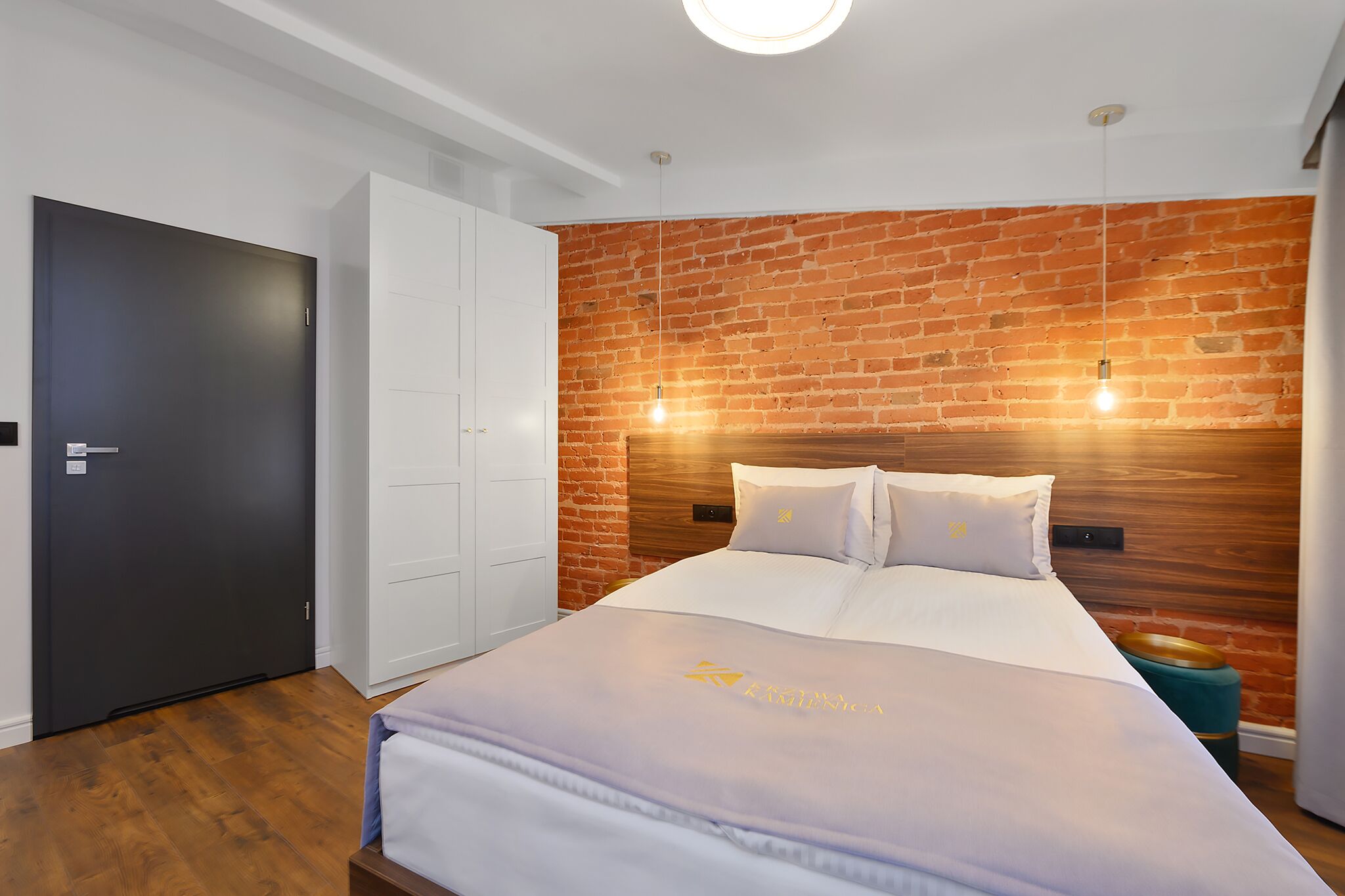 A modern bedroom with a brick wall, a large bed with white bedding, a wardrobe, and a door.