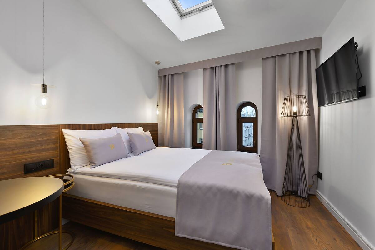 Ein modernes Hotelzimmer mit Oberlicht, einem Bett mit Kopfteil, einem Nachttisch, einer Stehlampe und einem an der Wand befestigten Fernseher.