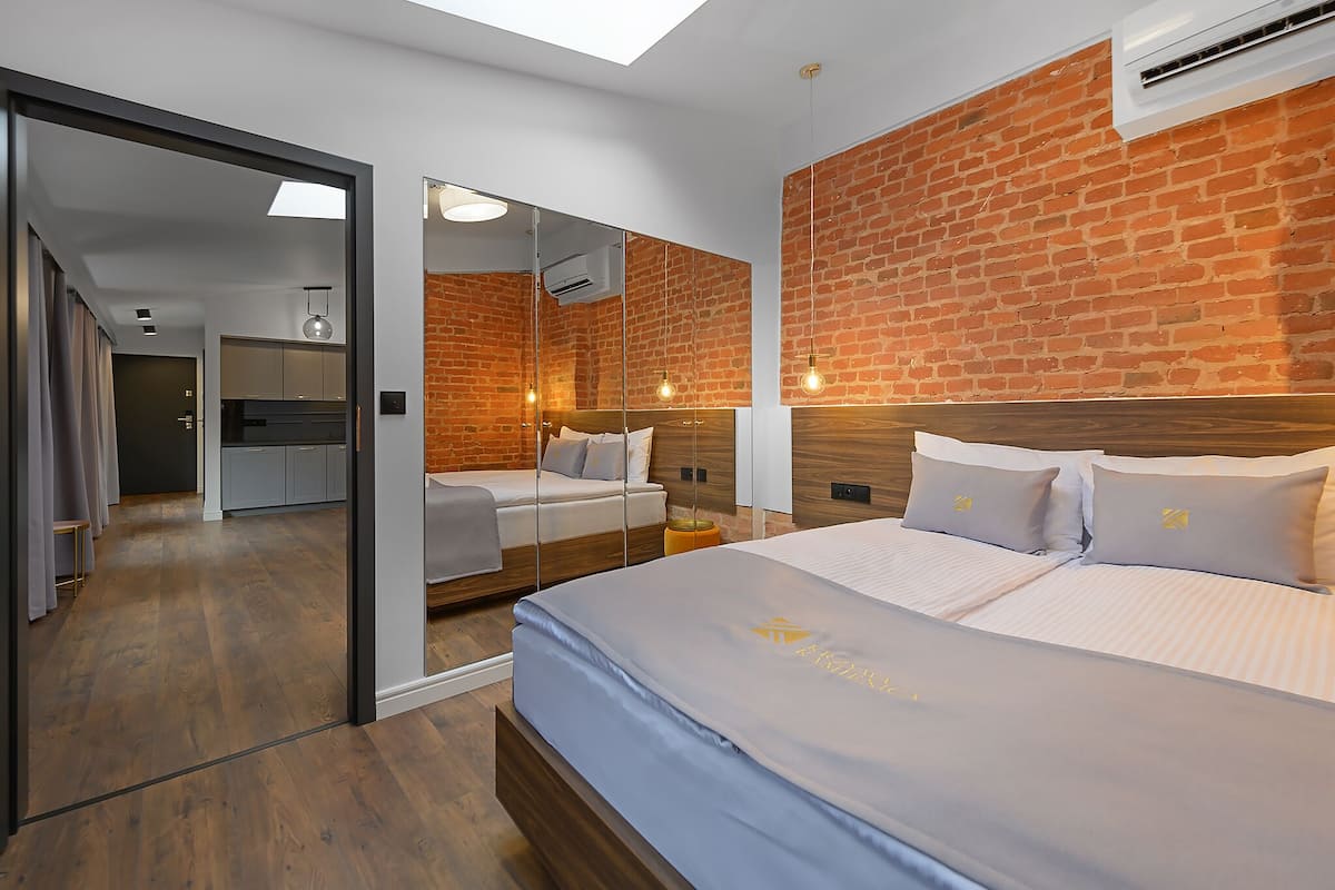 Ein modernes Hotelzimmer mit Backsteinwand, einem großen Bett mit Holz-Kopfschild, einem kleinen Nachttisch und einem Spiegel.