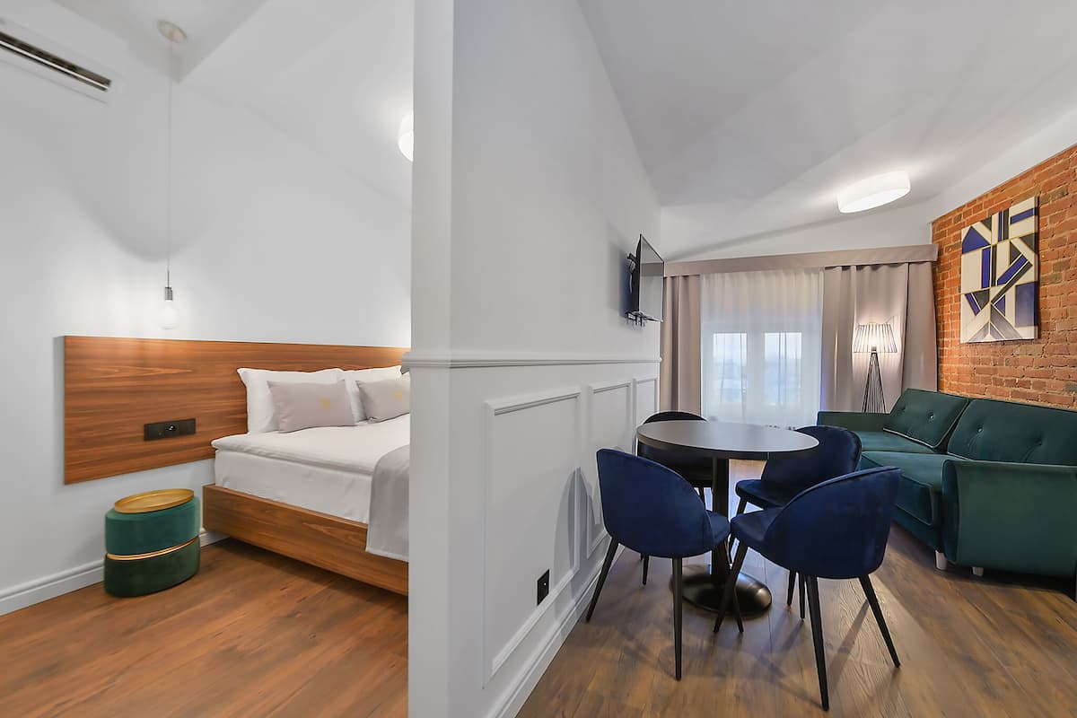 Ein modernes Hotelzimmer mit einem Bett mit Holz-Kopfstütze, einem grünen Sofa, einem runden Tisch mit blauen Stühlen und einer Ziegelwand mit einem geometrischen Gemälde.