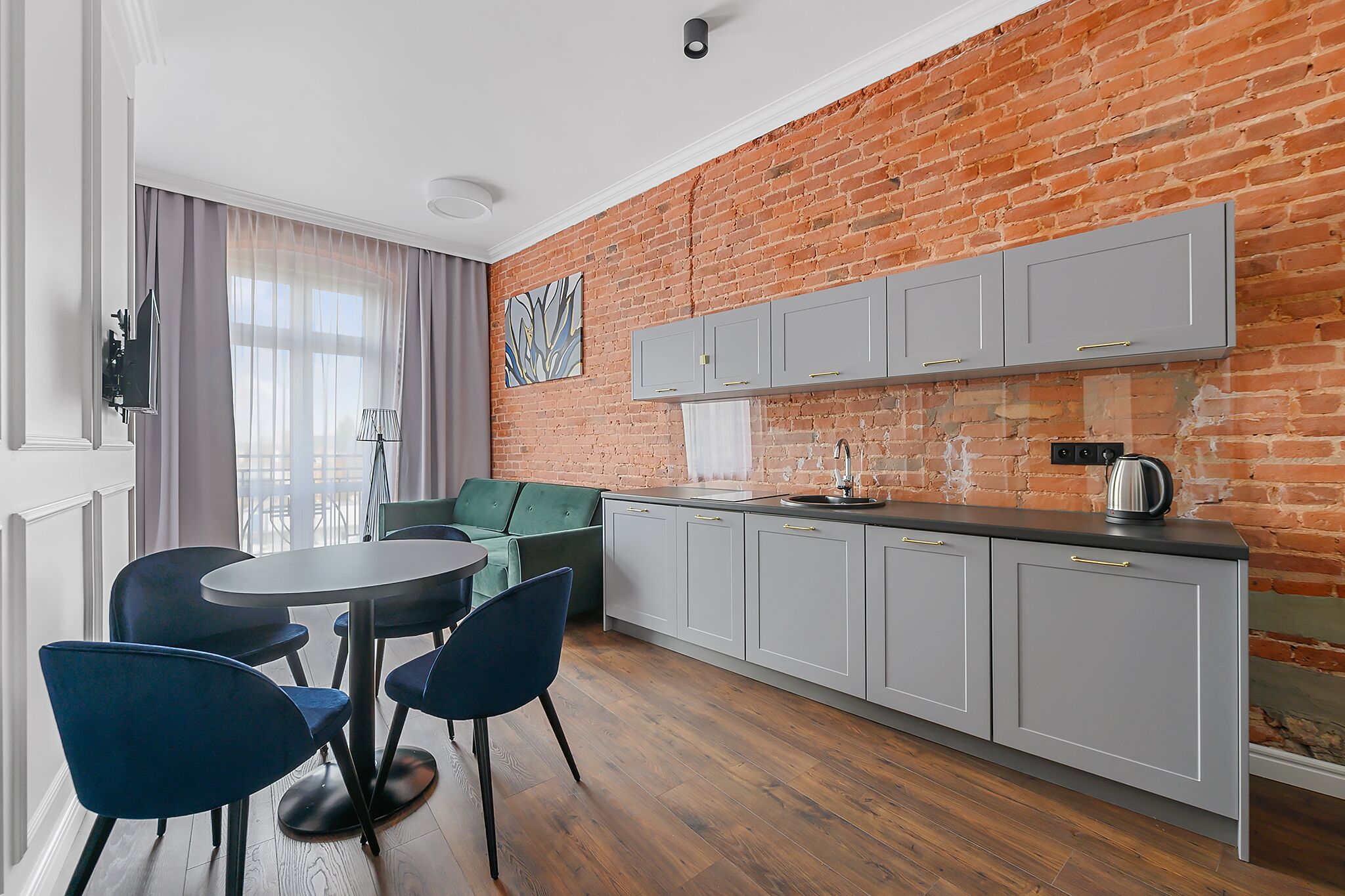 A modern kitchen with grey cabinets, a round dining table with blue chairs, and a brick wall.
