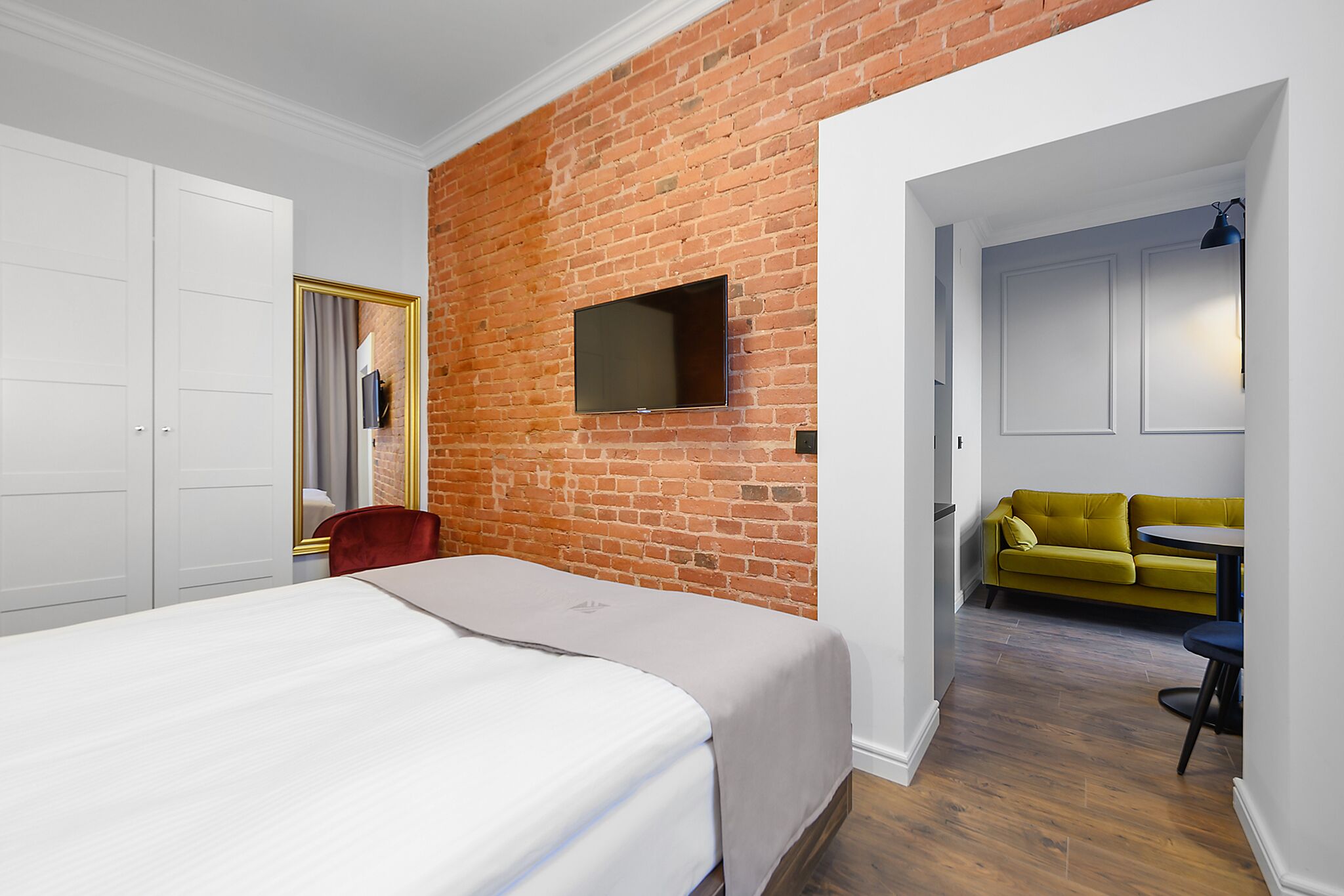 Una camera d'albergo con un muro in mattoni, una televisione a schermo piatto, un letto con copripiumino bianco, una poltrona rossa, un divano giallo e un piccolo tavolo con una lampada.