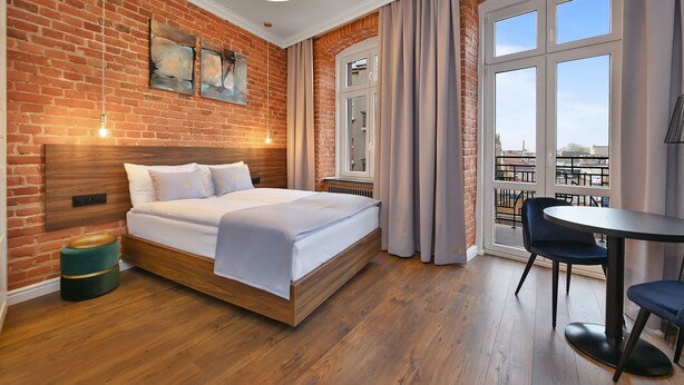 Ein Hotelzimmer mit Backsteinwand, einem Bett, einem Fenster mit Vorhängen, einem Tisch mit Stühlen und Kunstwerken an der Wand.