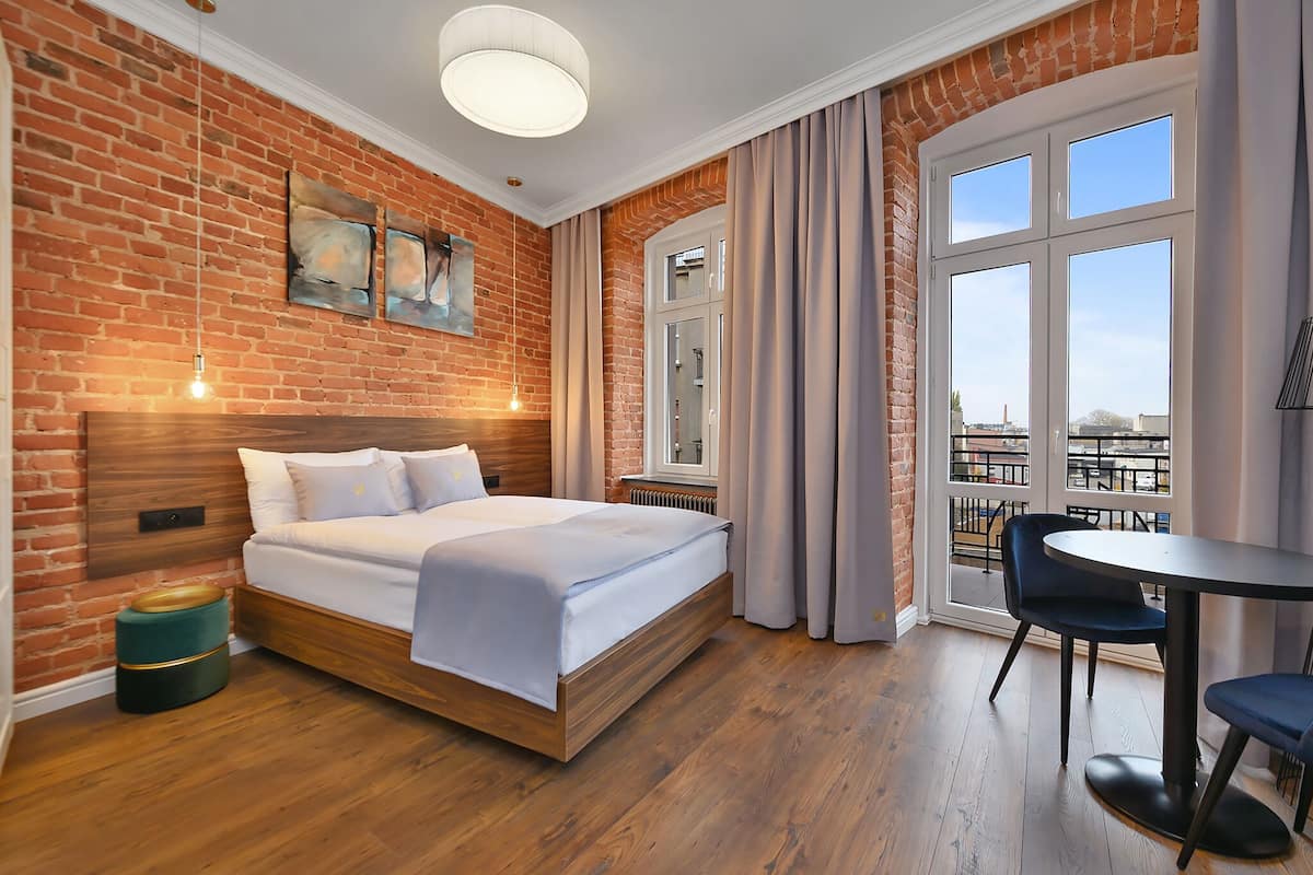 Ein Hotelzimmer mit Backsteinwand, einem Bett, einem Fenster mit Vorhängen, einem Tisch mit Stühlen und Kunstwerken an der Wand.