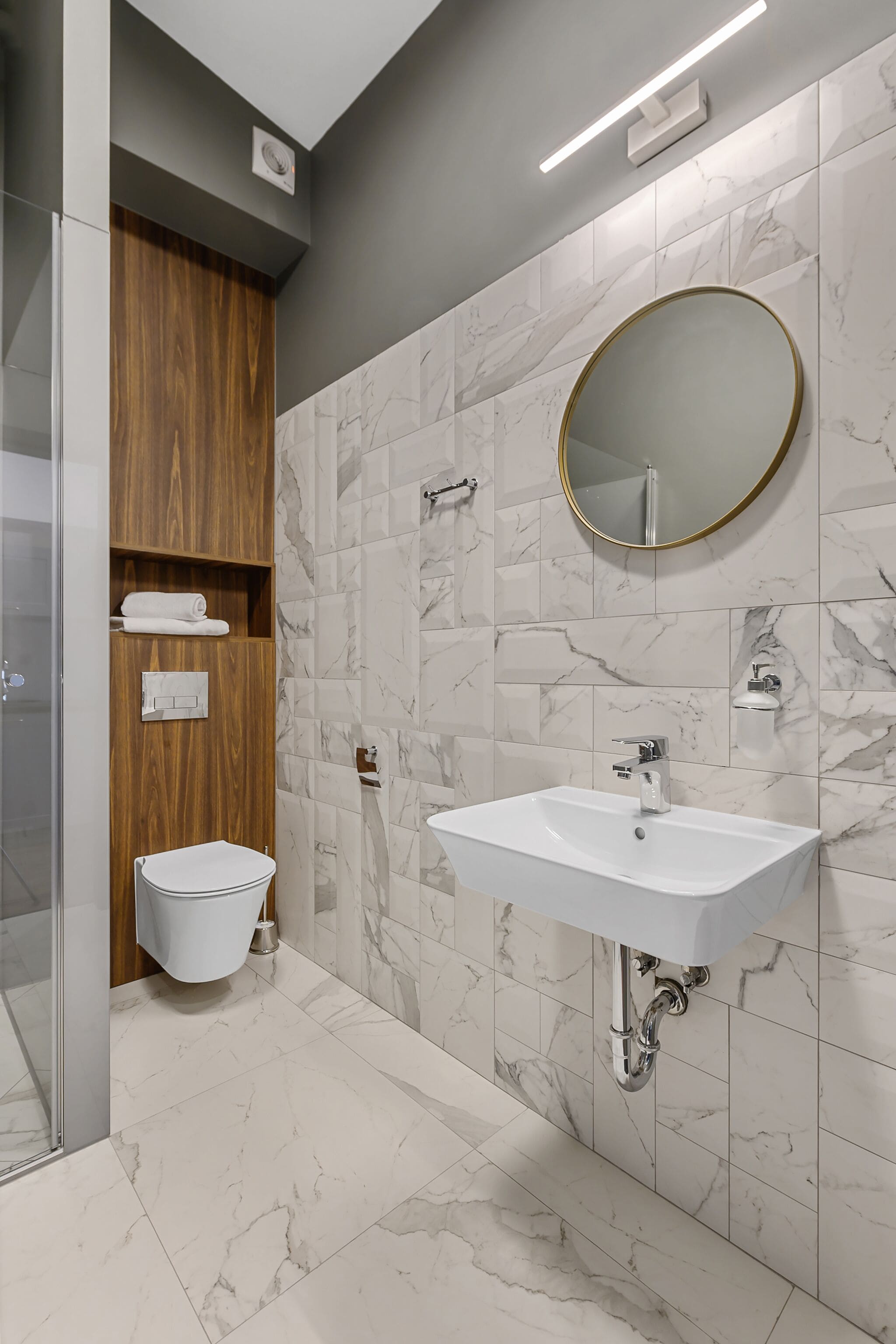 Un baño moderno con salpicadero de azulejos de mármol, tocador de madera, lavamanos blanco y espejo redondo.