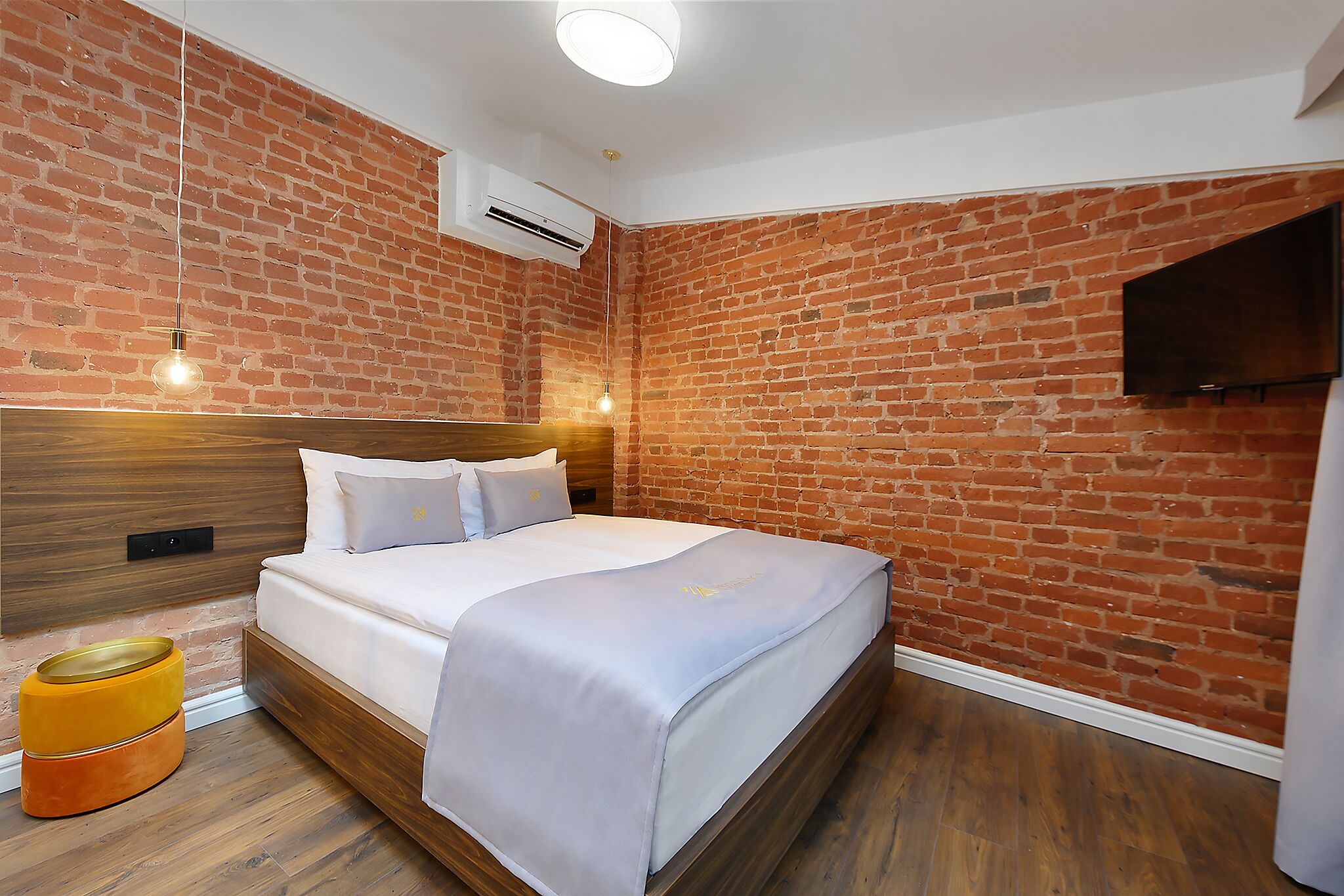 A modern hotel room with a brick wall, a wooden headboard, a bed with white bedding, a flat-screen TV mounted on the wall, and a wooden floor.